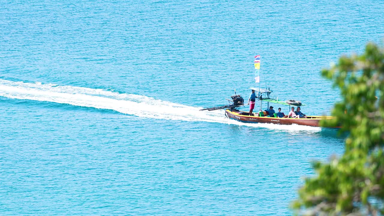 Longtail boat with passengers moves quickly across turquoise sea, viewed from above through foliage