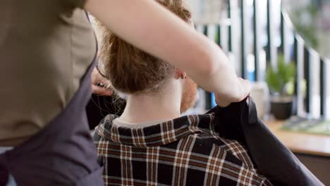 caucasian female barber putting cape on male client at barbershop, in slow motion