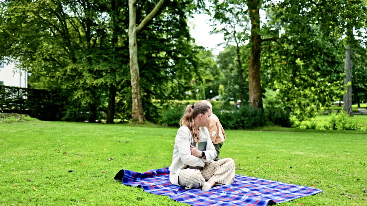 En ung mor sidder på et picnictæppe i en grøn park med sin lille søn. Den lille søn løber ved siden af sin mor og kigger interesseret på den bærbare computers skærm. Den unge mor arbejder fjernstyret.