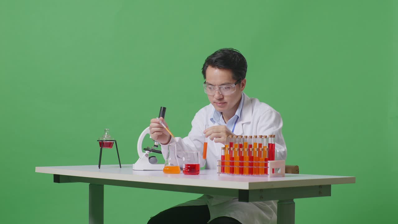 Side View Of Asian Man Scientist Sitting And Making Experiment With Test Tube On The Table With Microscope In The Green Screen Background Laboratory