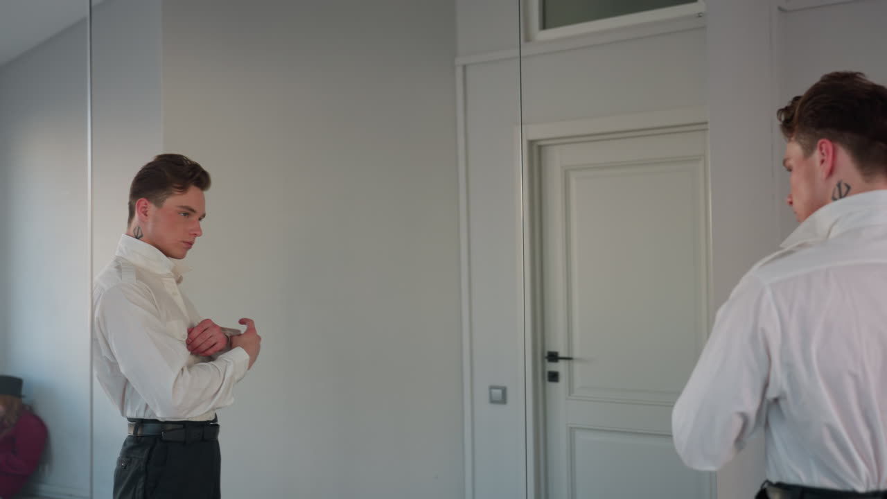 rear view man buttoning crisp white shirt adjusting belt while standing in front of full length mirror inside bright studio with reflective doors and minimalist loft background preparing for workday