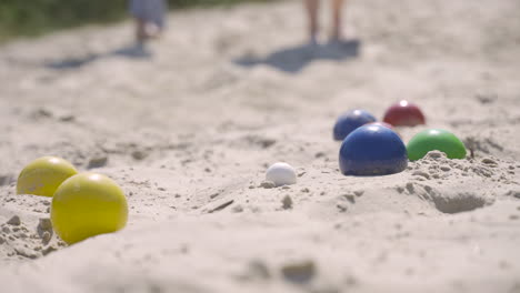 Close-up-view-of-colorful-petanque-balls-on-the-sand-on-the-beach,-then-a-player-throws-another-ball-nearby