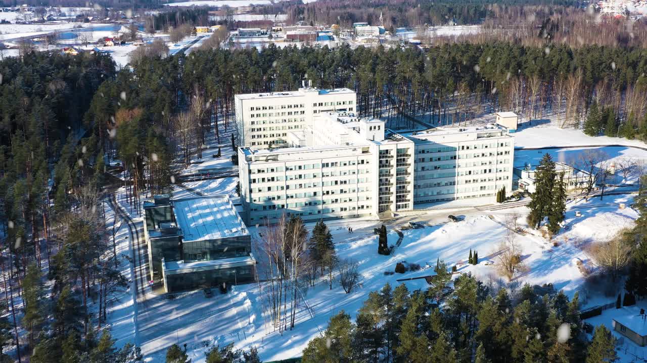 Sykehuskompleks i en skog om vinteren med fallende snø, medisinsk anlegg i et snørikt miljø, tilbyr helsetjenester og essensielle tjenester i fugleperspektiv.