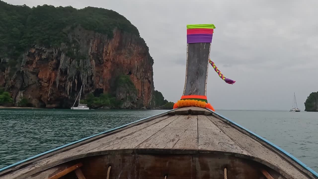 A vibrant longtail boat sails towards a towering, lush cliff under a cloudy sky.