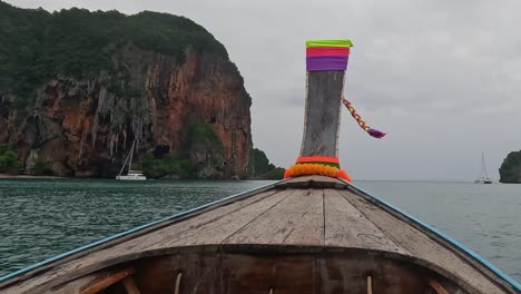 a vibrant longtail boat sails towards a towering, lush cliff under a cloudy sky.