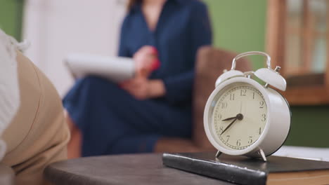 white retro style clock placed on thick cover notebook on table with blurred background of woman seated on sofa holding pen and writing pad during thoughtful session in calm room setting