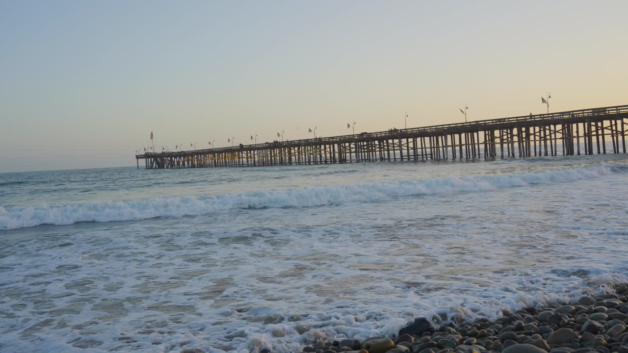 남부 캘리포니아에 위치한 백그라운드에서 부두와 일몰이 있는 벤추라 해변의 해안을 따라 부서지는 파도의 패닝 샷