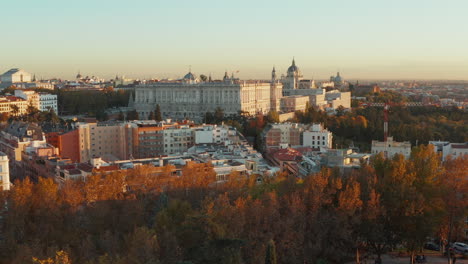 Tiefflug-über-Bäumen-Im-Park-Und-Gebäuden-Im-Stadtteil.-Blick-Auf-Den-Majestätischen-Königspalast,-Beleuchtet-Von-Der-Untergehenden-Sonne.