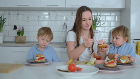 young beautiful mom in a white dress with two children are smiling and eating fresh burgers in their kitchen