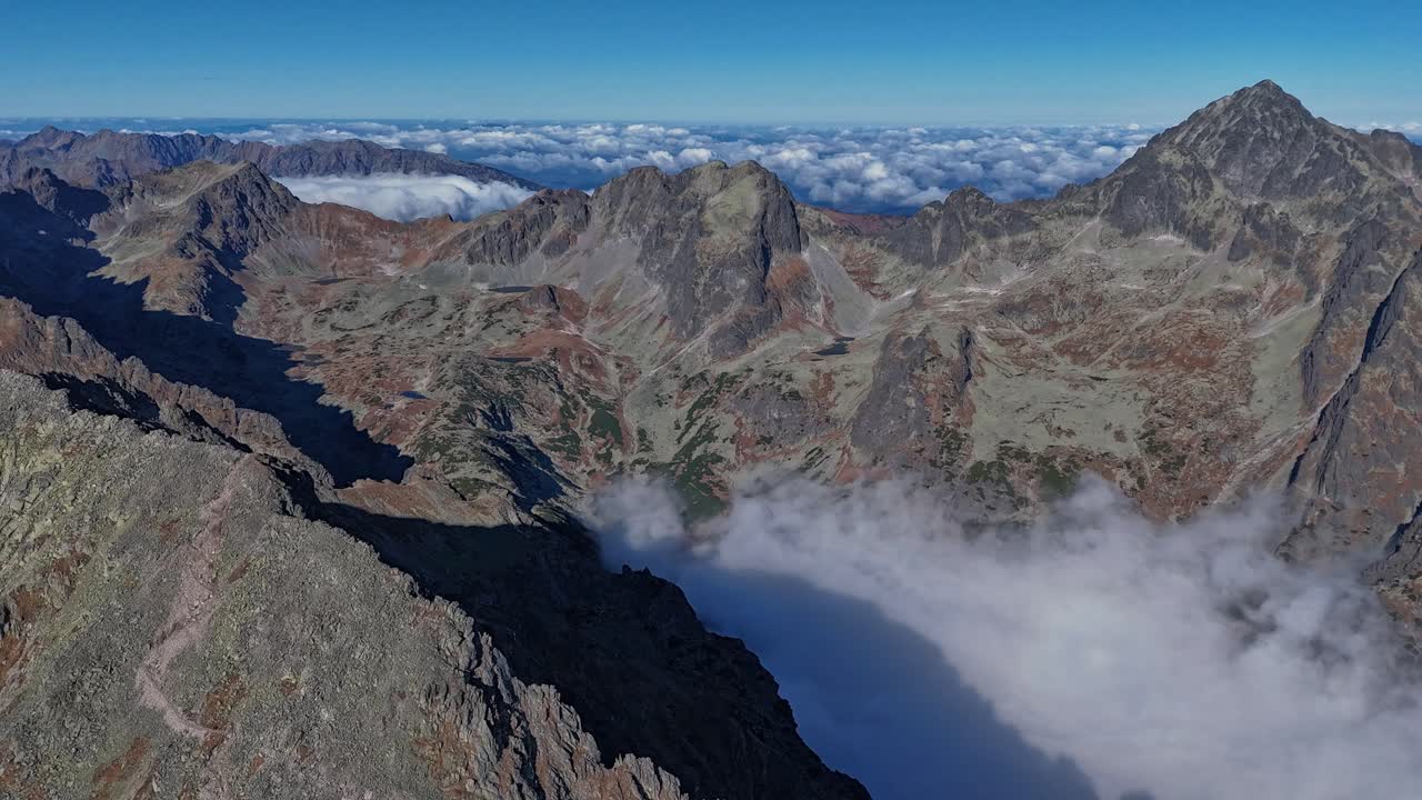 Hyperlapse drone shot over the High Tatras mountains, revealing rugged peaks and valleys blanketed in fog under a clear blue sky, creating a dramatic alpine scene. Location: High Tatras, Slovakia
