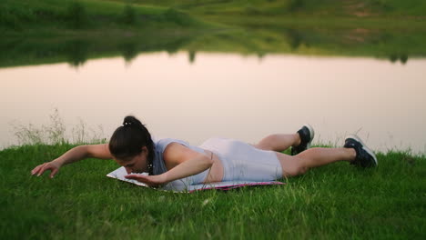 a rejuvenated woman in the background of a lake in a park lying on a gymnastic mat on her stomach performs a body lift to train her back muscles