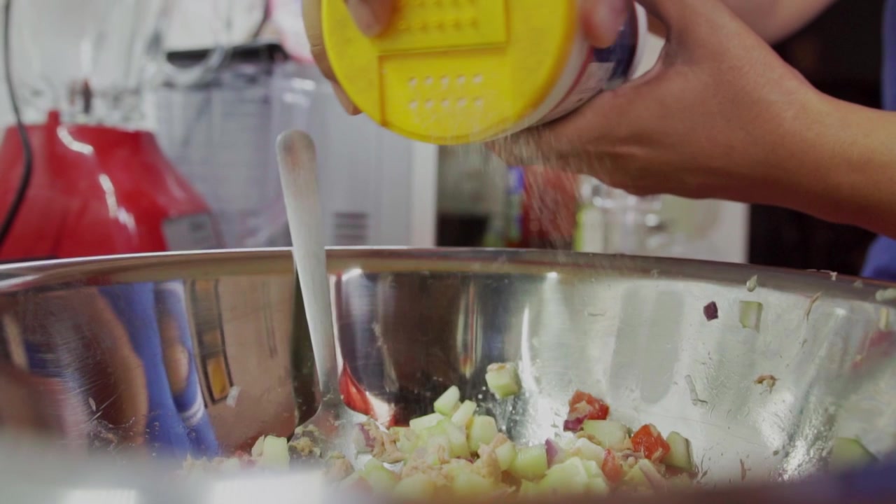 Primo piano delle mani di una donna che preparano ceviche di tonno con pesce a cubetti e verdure in una ciotola di metallo, mentre sparge sale in cucina.