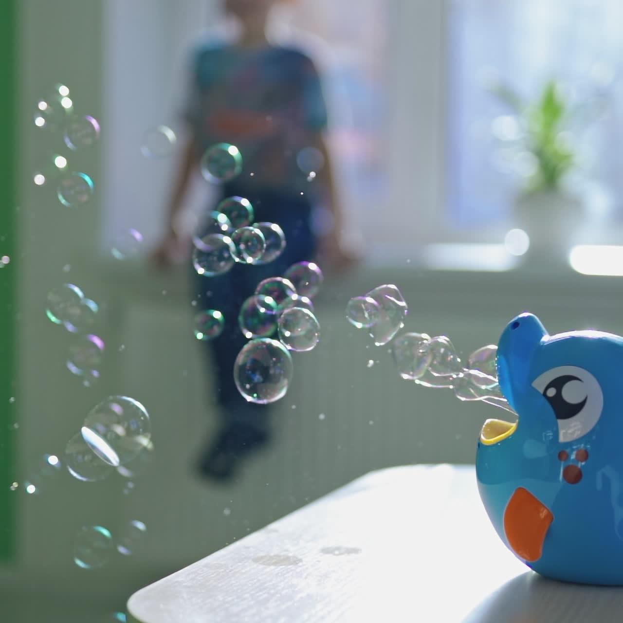 Many small bubbles scatter in one direction from the special appliance on the table indoors. Pretty device in a form of blue fish for creating bubbles on the blur background with a boy.