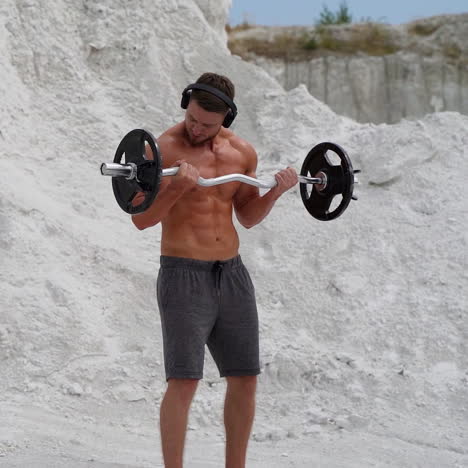 bodybuilder lifting barbell and looking on his muscular body outside. athlete man is training press muscles with metal barbell on white mountain background.