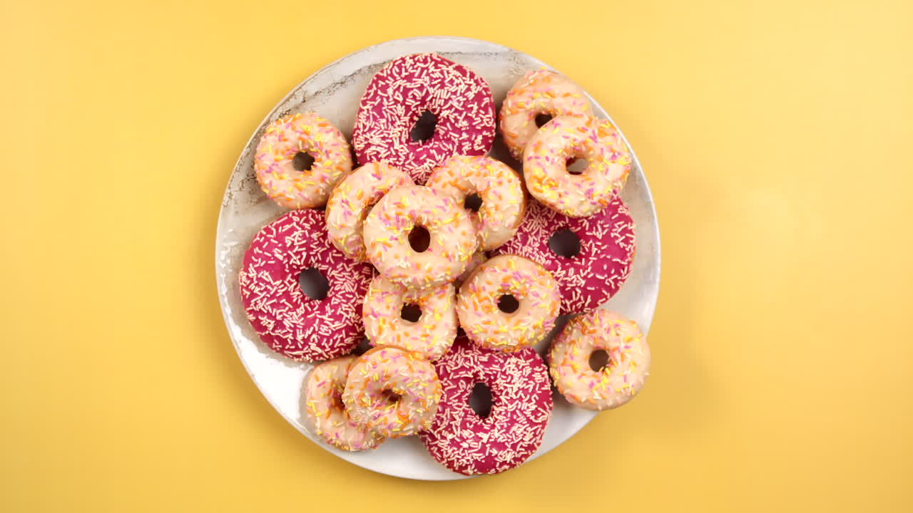 Plate full of colorful Donuts in different size, pink and white on light yellow background. Female Hand take small doughnut. Top view, filmed in Prores.