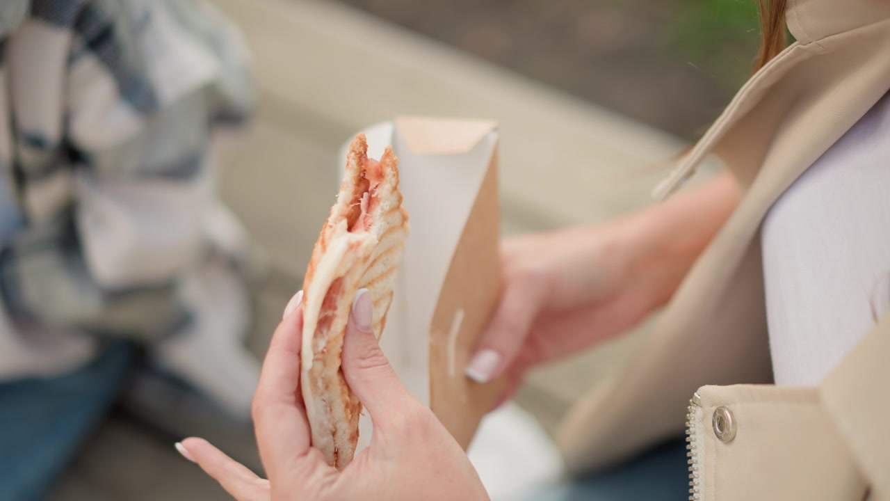 Close up of woman holding and eating open faced sandwich with visible grill marks while another hand holds paper container. scene is set outdoors on pavement with soft natural light