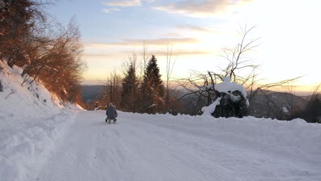 female on sled carrying a baby slowly descends down snowy road into golden sunset