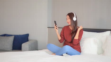 a young woman sitting on comfy bed dances to the music from her smartphone and headphones