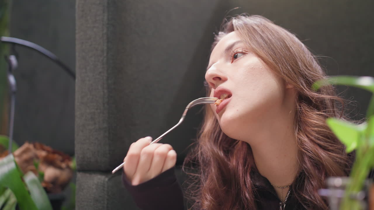 Lady with brown curly hair holds fork with waffle, staring at it thoughtfully before eating slowly, surrounded by cozy booth and greenery in relaxed, softly lit environment with subtle decoration