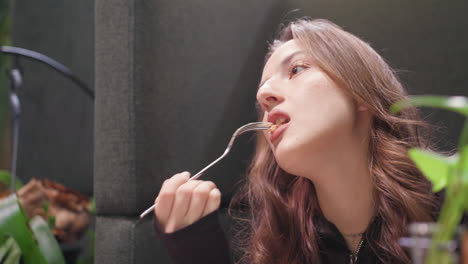 lady with brown curly hair holds fork with waffle, staring at it thoughtfully before eating slowly, surrounded by cozy booth and greenery in relaxed, softly lit environment with subtle decoration
