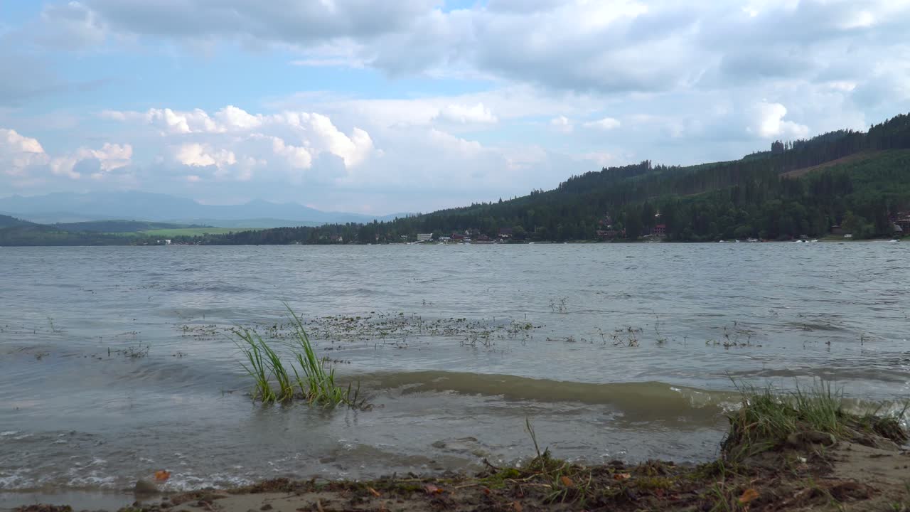 작은 파도가 호수를 강타하고 보트, 언덕, 산과 함께 멀리 이동하는 구름의 빠르게 움직이는 낮은 각도 시간 경과