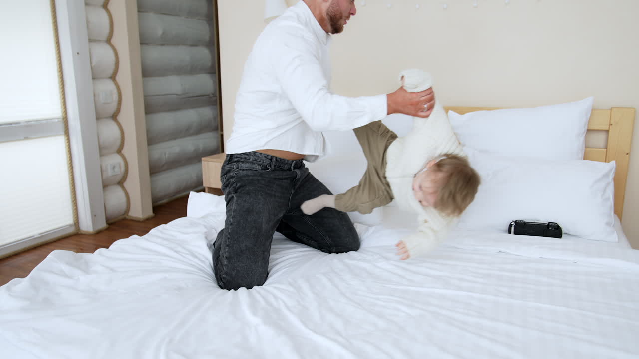 Smiling bearded Caucasian father wearing white shirt plays with his baby. Man throws a son on the bed and pillows playfully.