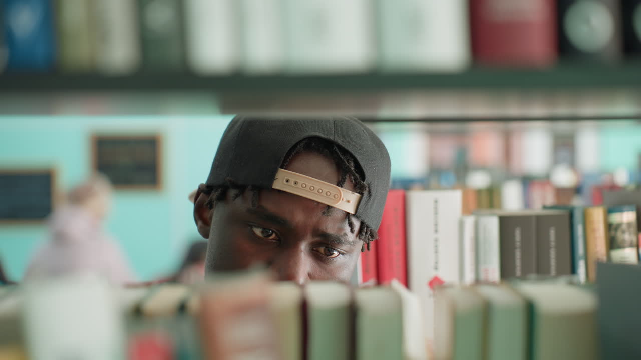 close up in library shows young man peering between shelves, scanning books row, fingertips grazing spines while eyes track titles, focused search mood, soft bokeh background