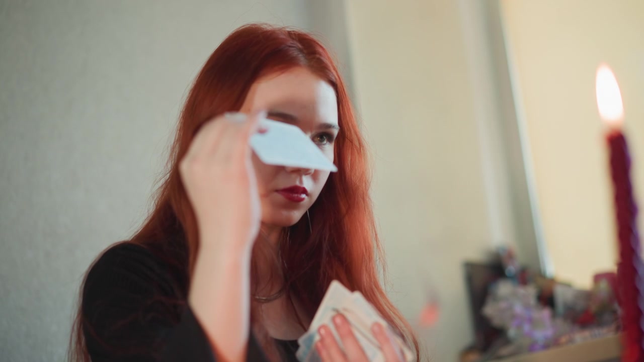 Redhead caucasian fortune teller reading cards by candlelight, intimate indoor scene with single tall candle, warm glow on face, contemplative expression, slow reveal of card, vintage table