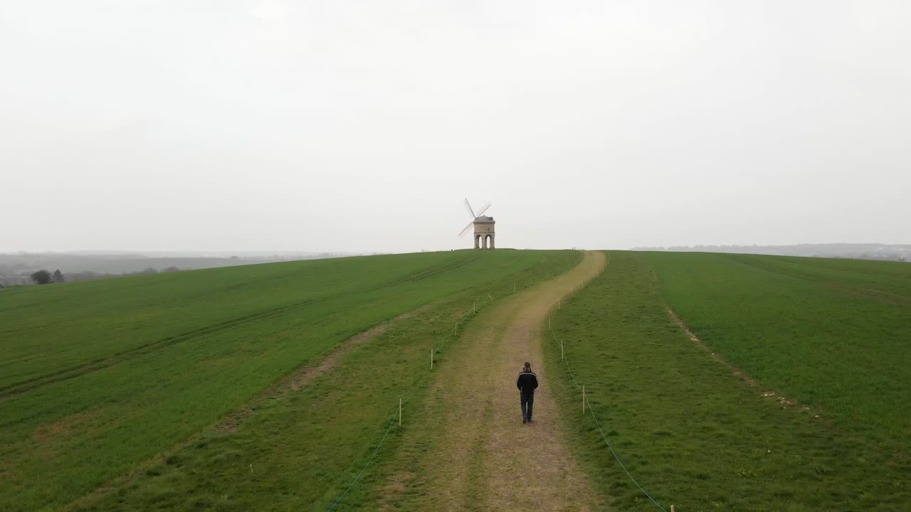 Aerial shot following a man walking along a path toward an old windmill on a hill.