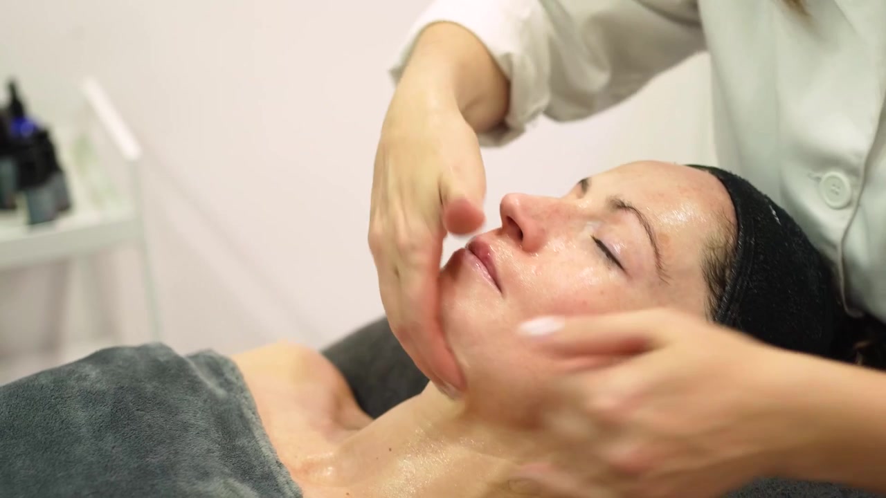 En kosmetolog masserer en kvindelig klient i ansigtet med en kosmetisk maske i en skønhedssalon.