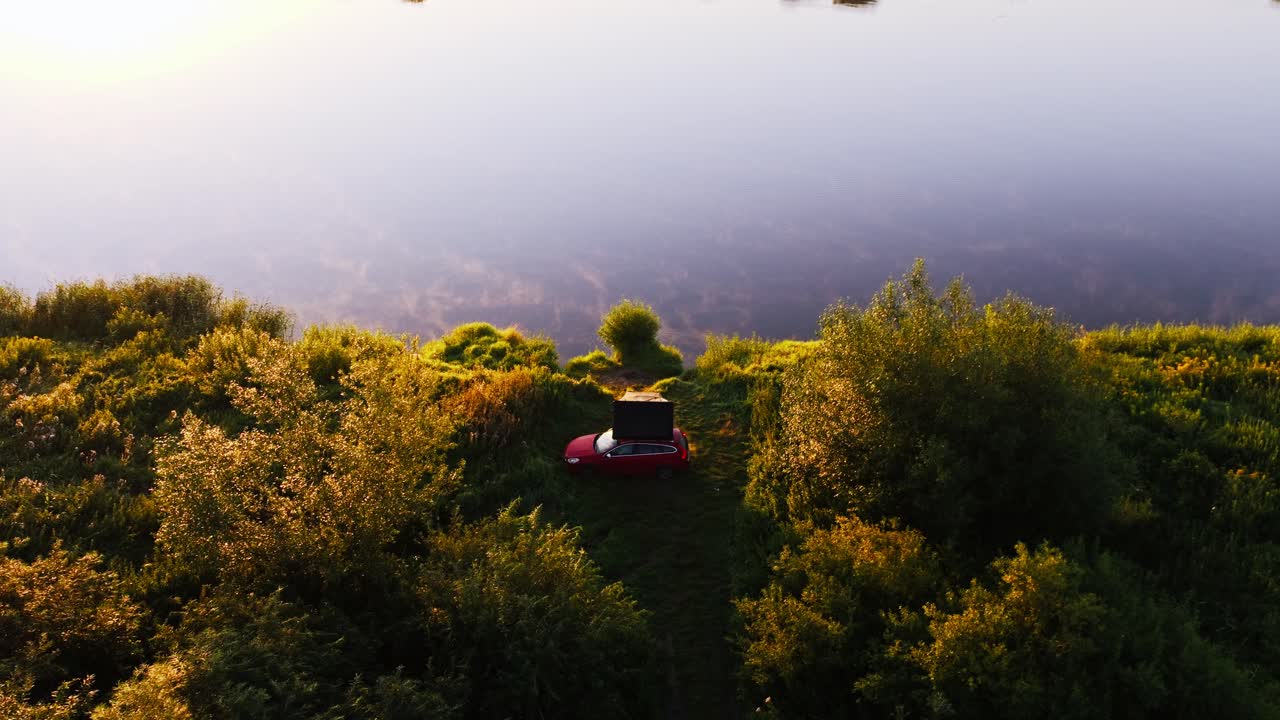 Drone turns left, ascends as golden hour paints red car, rooftop tent by Vistula