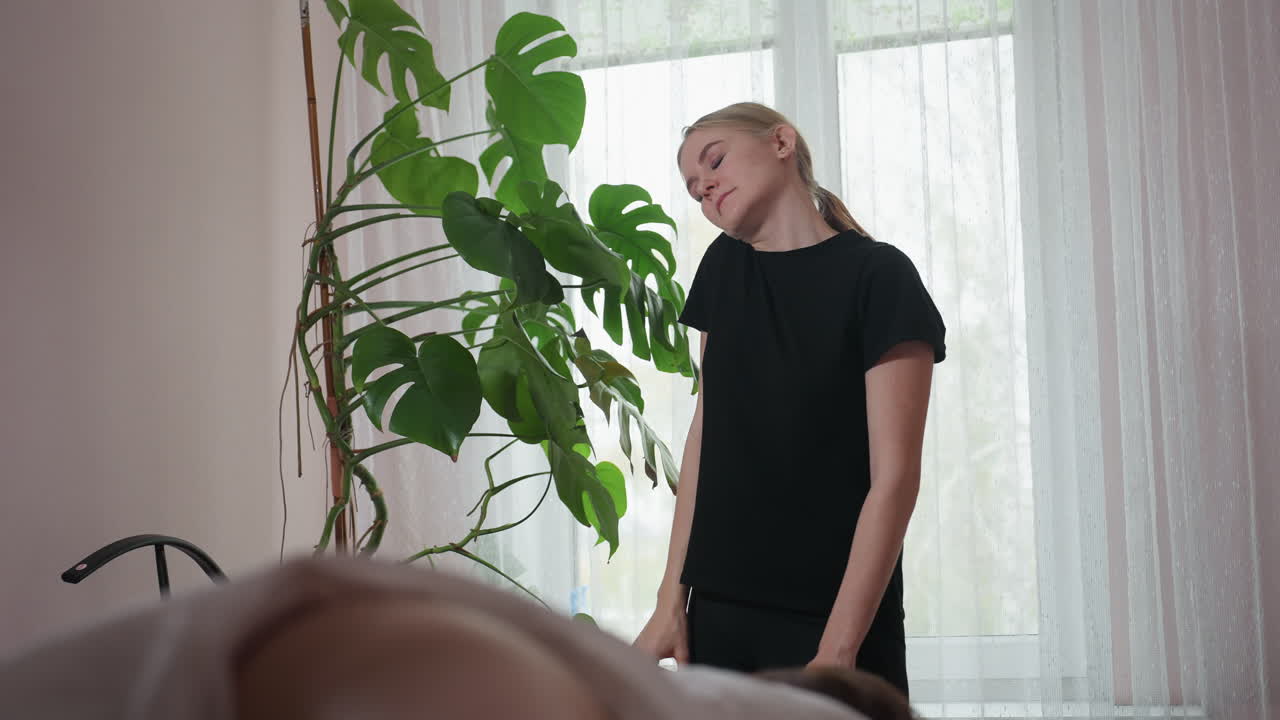 Massage therapist standing near leafy plant, stretching neck upward in moment of fatigue during wellness session, sunlight filtering through sheer curtains creating serene and calm indoor atmosphere
