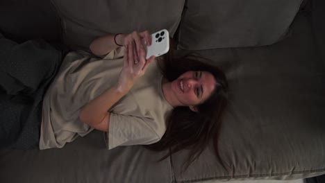 From-above-close-up-of-a-happy-brunette-girl-in-a-beige-T-shirt-lies-on-a-gray-sofa-and-looks-at-her-white-smartphone-watching-a-video.-Happy-girl-relaxing-and-lying-down-on-the-sofa-with-her-smartphone