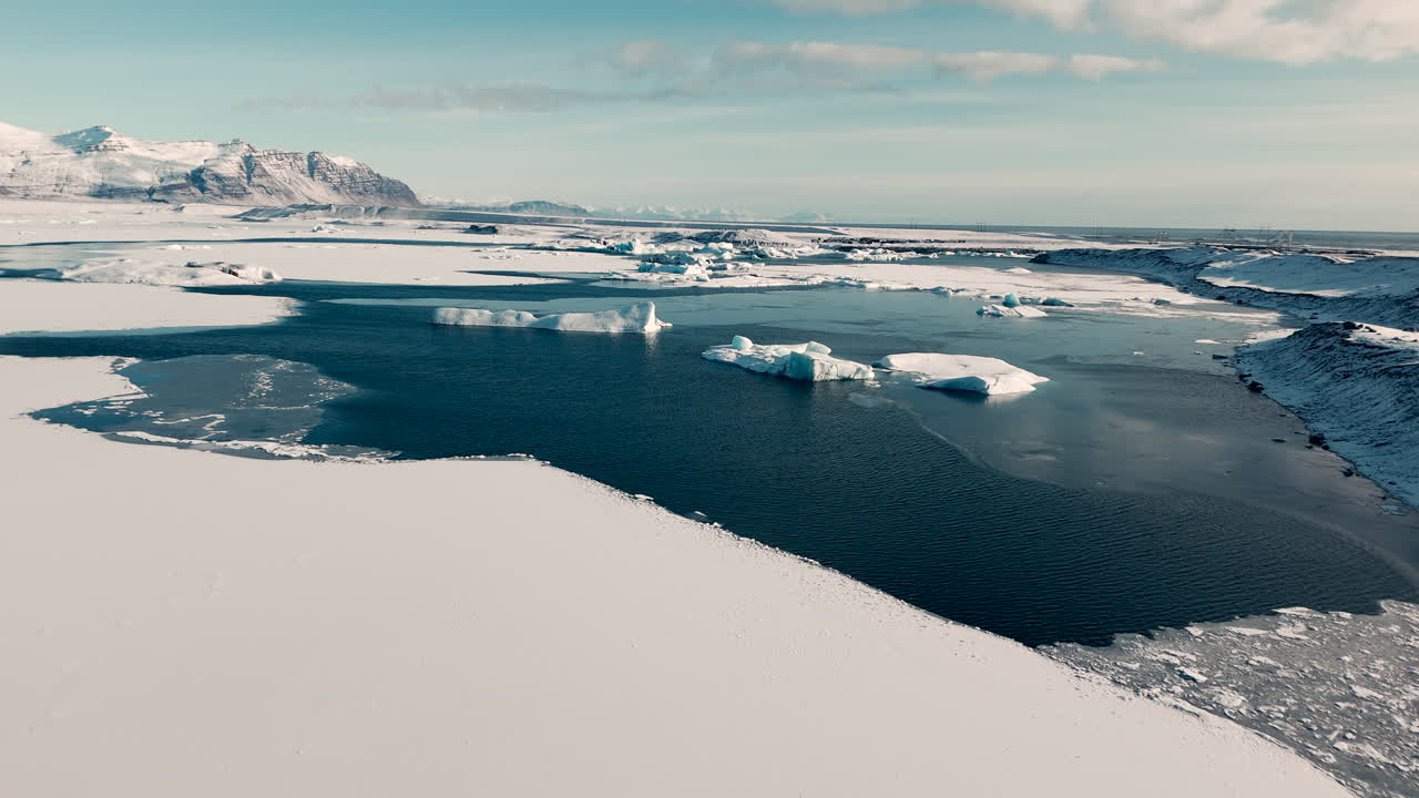 İzlanda'daki Jökulsárlón'da, karlı bir manzara üzerinde, okyanusta buzdağları ve arka planda dağların yer aldığı drone çekimi.