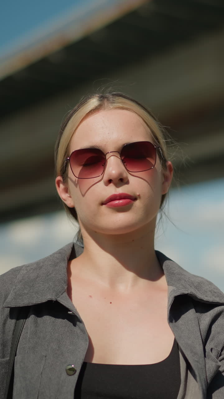 회색 셔츠와 선글라스를 입은 백인 여성이 여러 가지 다채로운 쇼핑 가방을 뒤집고 야외에 서 있습니다.그녀는 배경이 약간 흐릿하고 다리를 특징으로하면서 자신감 있게 서 있습니다.