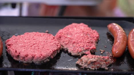 Sausages-and-under-un-cooked-raw-rare-red-beef-hamburger-patties-frying-sizzling-splattering-on-cast-iron-skillet-grill-at-campsite-barbecue-during-the-summertime