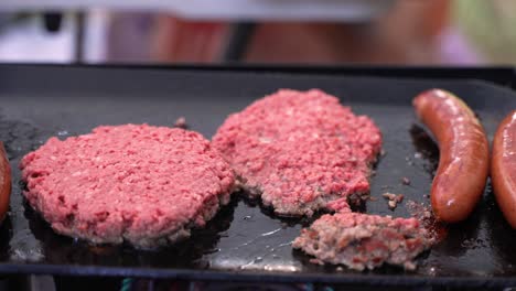 sausages and under un-cooked raw rare red beef hamburger patties frying sizzling splattering on cast iron skillet grill at campsite barbecue during the summertime