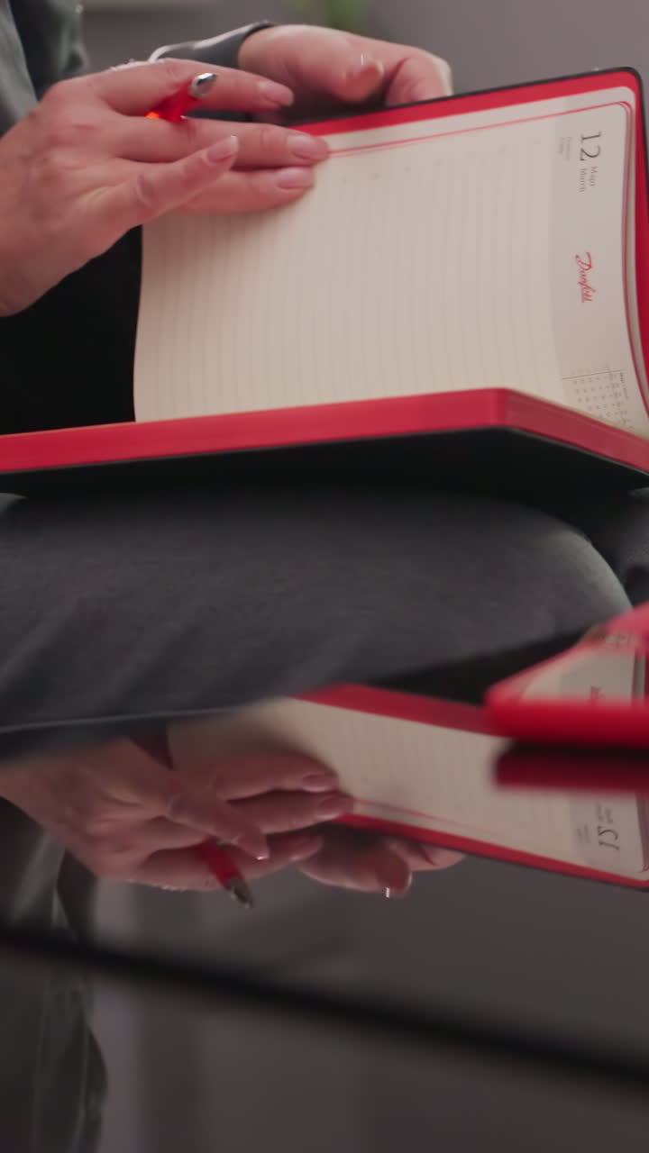 Close-up of businesswoman's hand resting on stack of red notebooks, smartphone, and papers on glass table. Female professional organized and focused, showcasing work environment and productivity