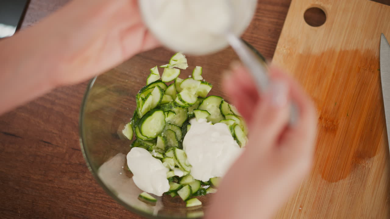aerial view of transparent glass bowl filled with sliced cucumber and white salad cream beside sharp knife on wooden board as person stirs ingredients gently in bright kitchen environment