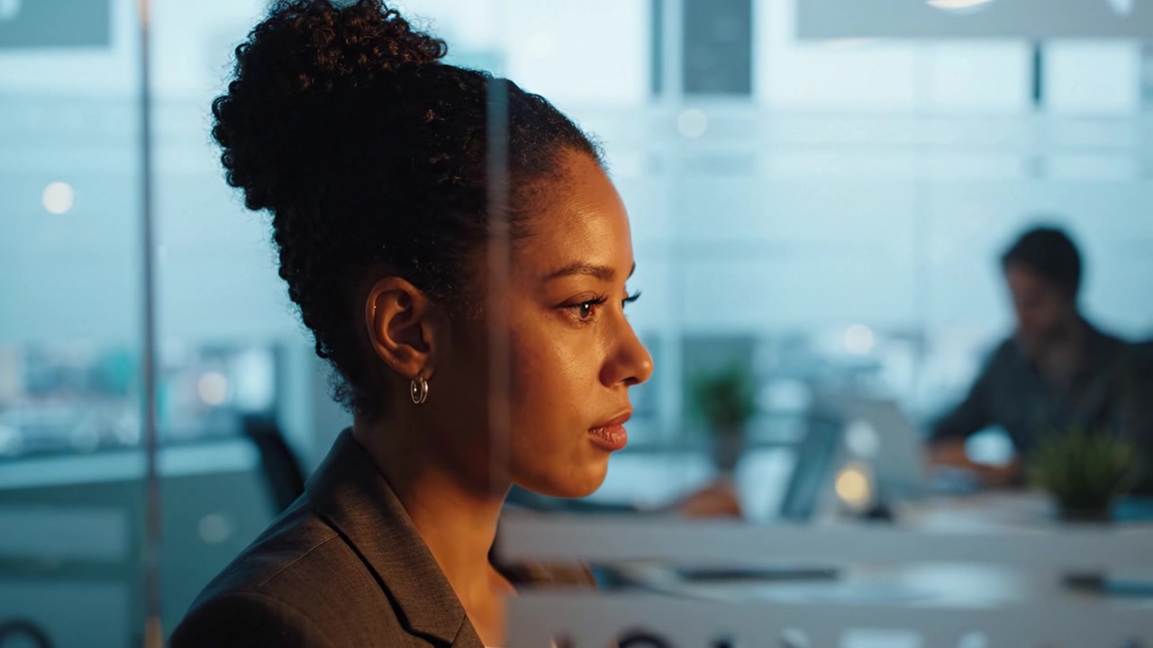 Focused Professional in Modern Office Environment Reflecting Thoughtfully While Colleagues Work in Background, Illuminated by Evening Light