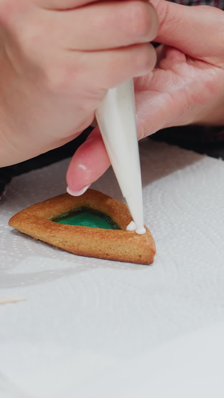 Close-up of baker using white piping bag to decorate cookie with green filling on tissue paper, toothpick nearby for precision as baker carefully applies icing to cookie edges