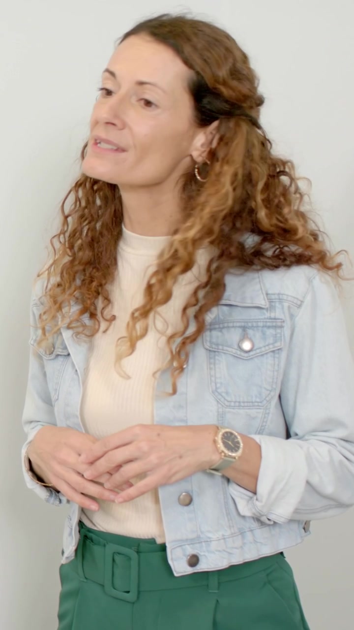 Video verticale: Donna con giacca di jeans che risponde, gesticolando verso una voce fuori campo vicino a un muro, mostrando la cintura.