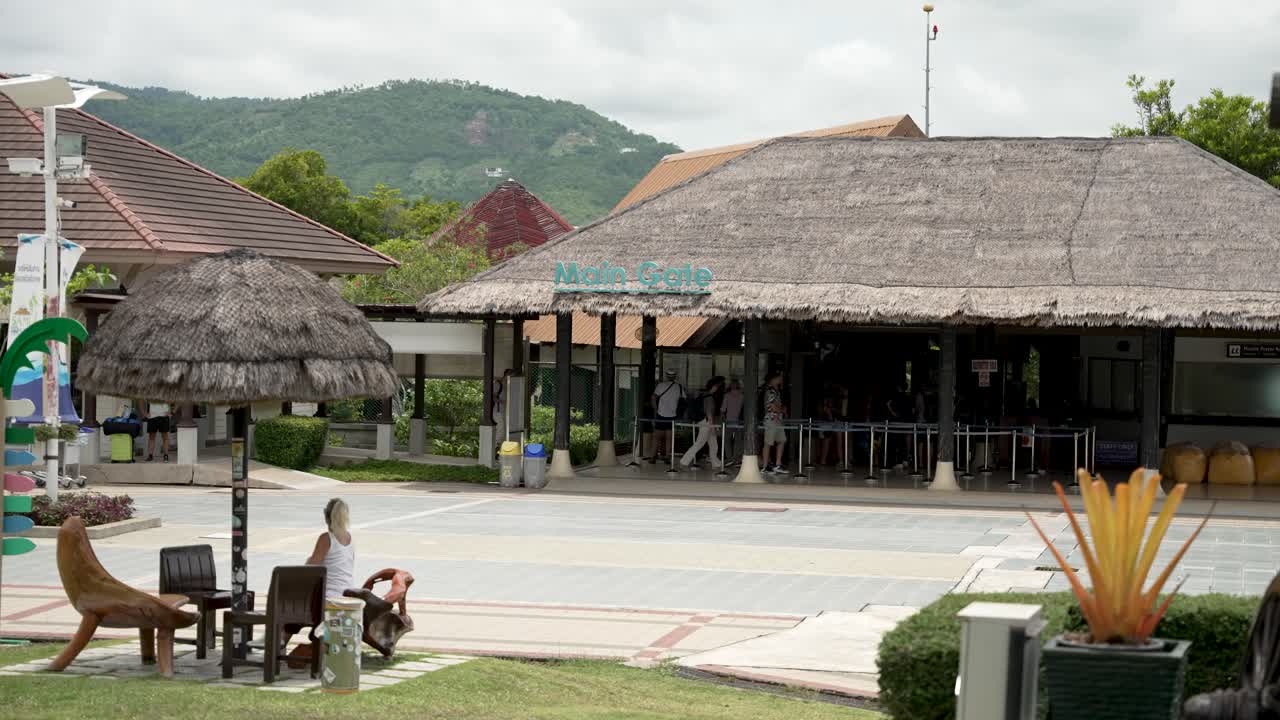 Scenic view of Koh Samui Airport's main gate set in a tropical outdoor environment. Features thatched roofs and people in a casual waiting area.