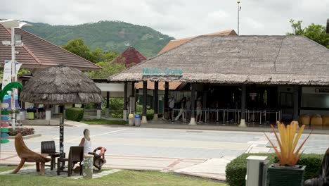 scenic view of koh samui airport's main gate set in a tropical outdoor environment. features thatched roofs and people in a casual waiting area.