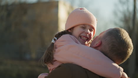 a joyful moment as a little girl in a pink cap and jacket is carried by a man in a brown jacket. she hugs him tightly with a big smile on her face, radiating happiness and warmth in an outdoor setting