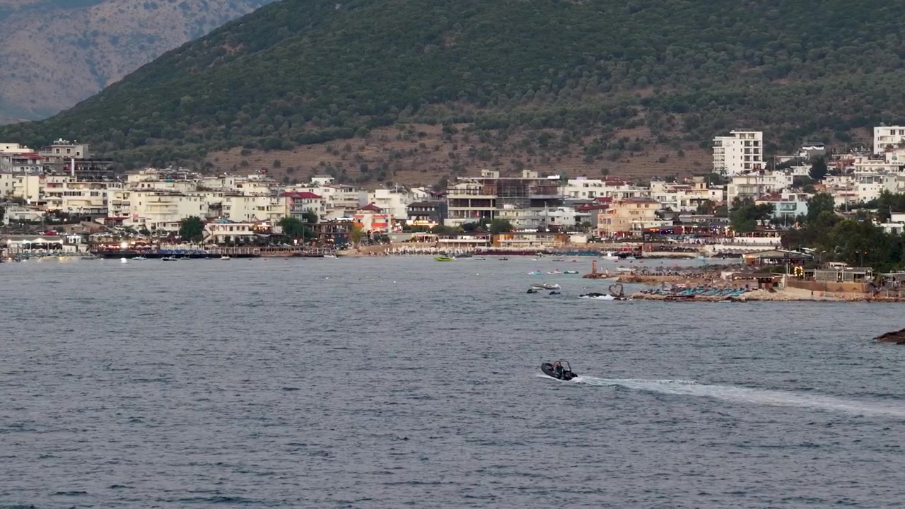 Arnavutluk'un Ksamil kıyısında seyreden sürat teknesi. Kıyı kasabası binalarının, yeşil tepelerin ve deniz suyunun manzarası. Akdeniz yaz tatili ve deniz yolculuğu.