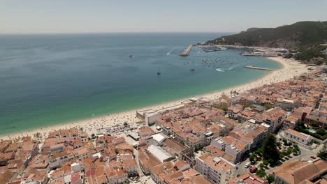 Vista-Aérea-Sesimbra-Hermosa-Costa,-Edificios-De-Tejas-Rojas-Frente-Al-Mar,-Playa-De-Agua-Clara