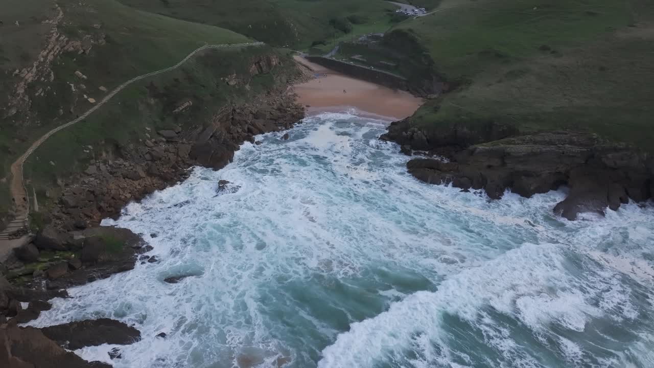 A drone flies at mid-altitude over the choppy waters of the Cantabrian Sea, approaching Santa Justa beach. The 16th-century hermitage stands atop the cliff, with hikers in the distance on the path