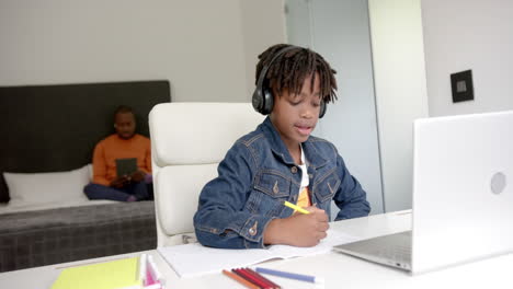 african american boy using laptop for online lesson with father in background at home, slow motion