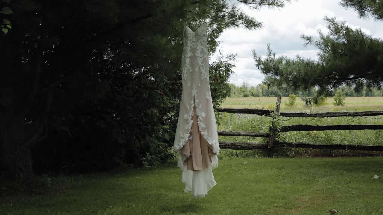 Elegancka, designerska biała suknia ślubna wisząca na sośnie na podwórku rozległej działki z drzewami, drewnianym płotem i polem uprawnym w tle, w Strathmere Wedding and Event Centre